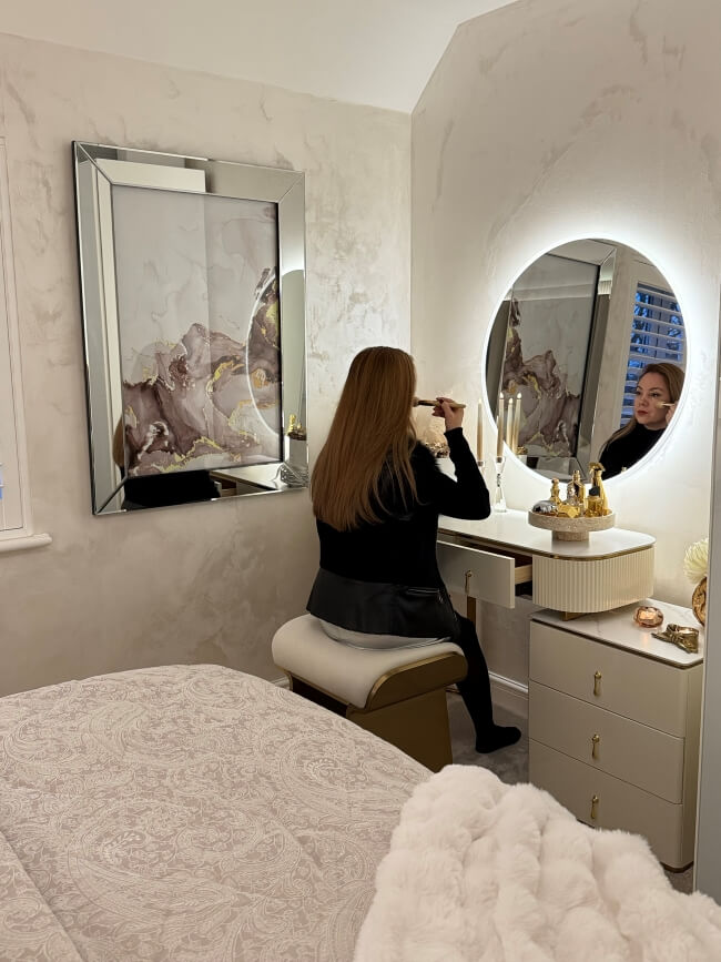 Woman applying makeup in front of a round mirror in a bedroom.