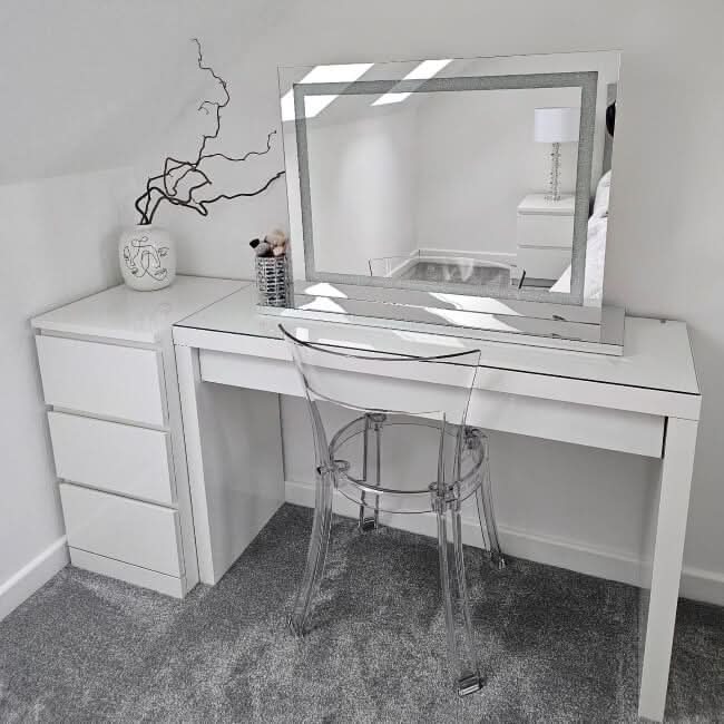 A modern vanity setup with the hollywood mirrors Hollywood Crystal Pro Mirror 80x60cm, featuring dimmable lights, a clear acrylic chair, small white drawers, minimalist decor with a vase and white piece, plus grey carpet and white walls.