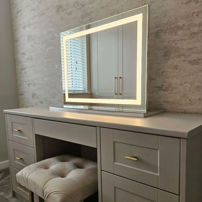The Hollywood Crystal Pro Mirror 80x60cm by hollywood mirrors, a modern vanity table set with gold handles, cushioned stool, and dimmable square mirror, is shown against a textured beige wall near a window with white blinds.