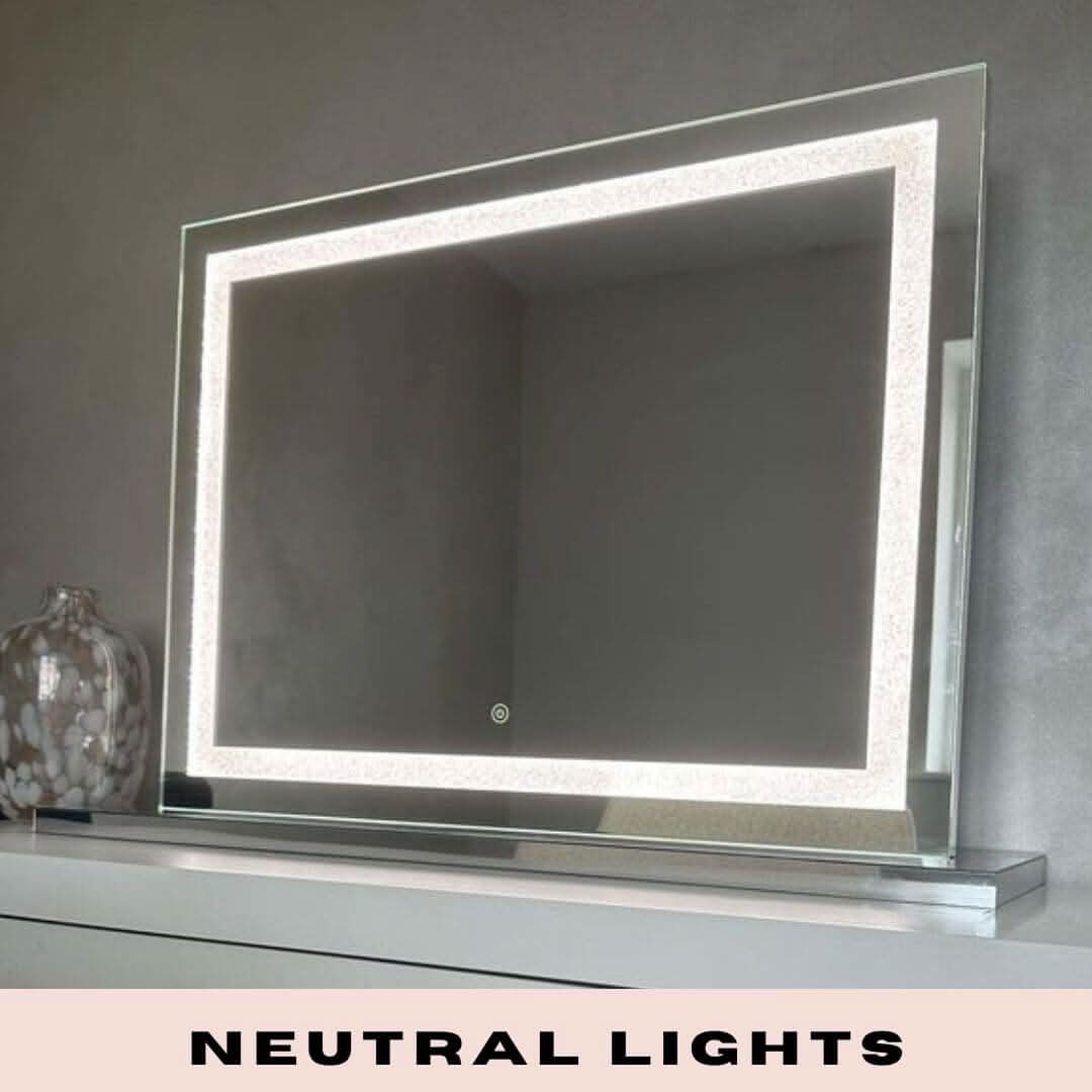 The hollywood mirrors Hollywood Crystal Pro Mirror 80x60cm, featuring built-in dimmable neutral LED strips along its border, sits on a white surface beside a decorative glass vase. NEUTRAL LIGHTS highlights its adjustable lighting settings.