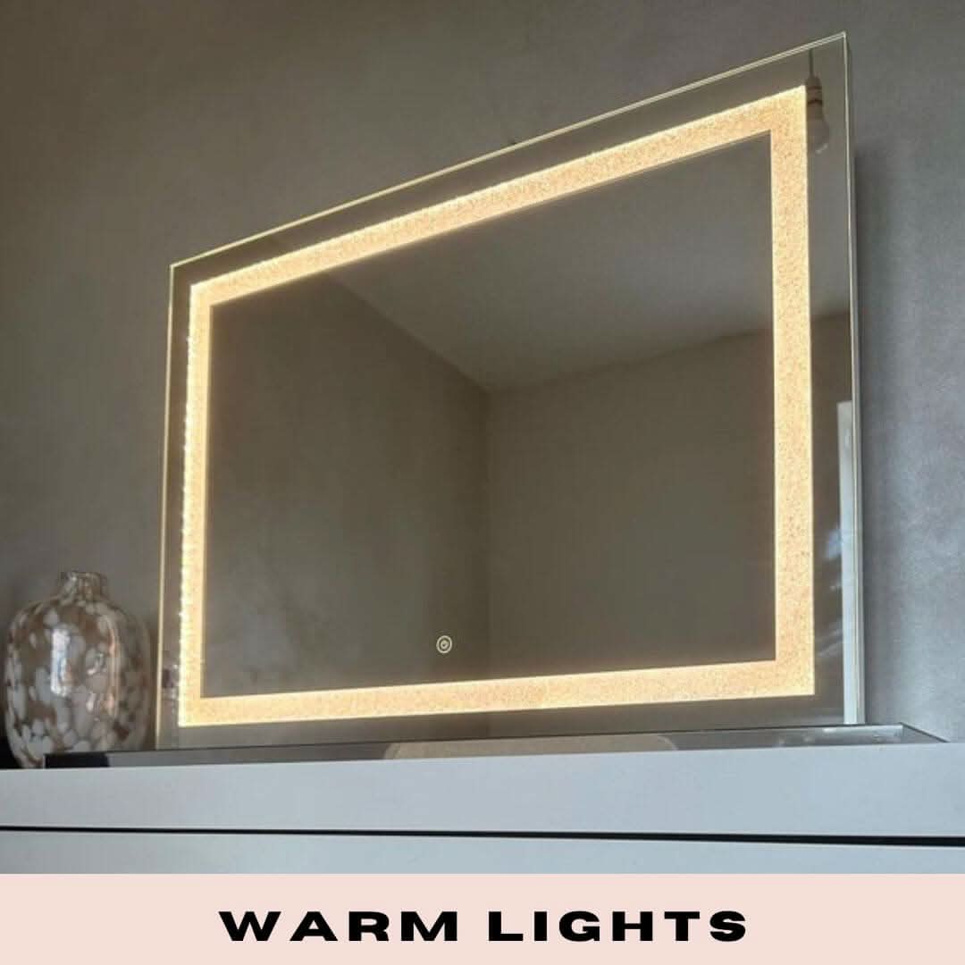 The Hollywood Crystal Pro Mirror 80x60cm by hollywood mirrors features a built-in warm LED strips border and is shown wall-mounted above a white surface with a decorative vase. WARM LIGHTS appears at the bottom of the image.