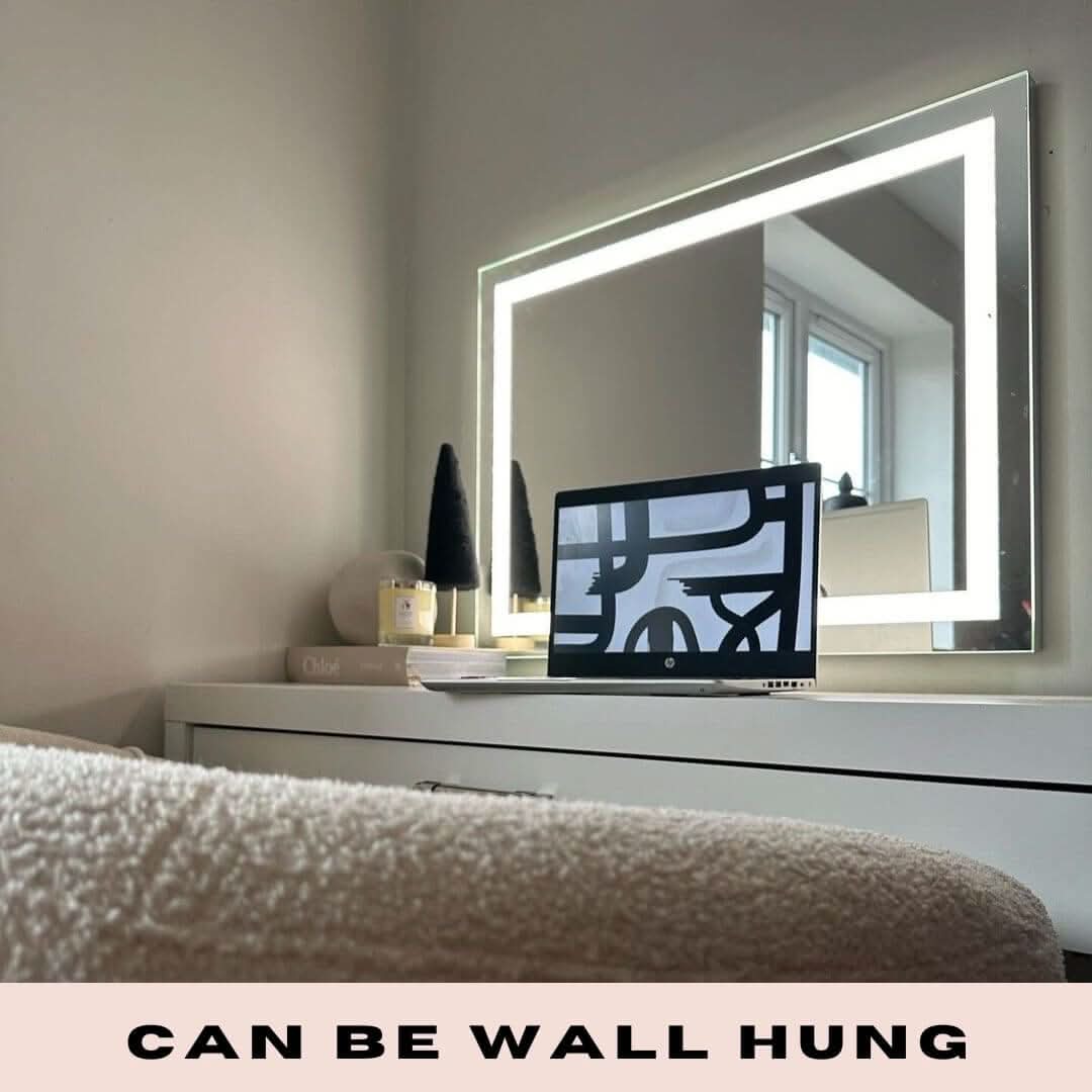 The Hollywood Crystal Pro Mirror 80x60cm by hollywood mirrors, a modern illuminated rectangular mirror with dimmable settings, is wall-mounted above a white dresser with a laptop, decor pieces, and two black cone-shaped ornaments. Text reads CAN BE WALL HUNG.