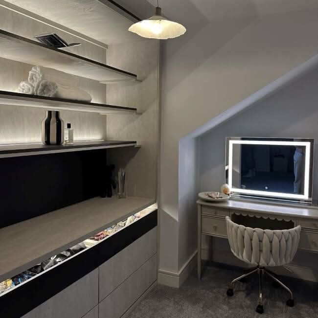 A modern, well-lit dressing room features open shelves for towels and bottles. Dimmable LED strips illuminate a vanity with the hollywood mirrors Hollywood Crystal Pro Mirror 80x60cm and a cushioned chair beneath a sloped ceiling by the window.