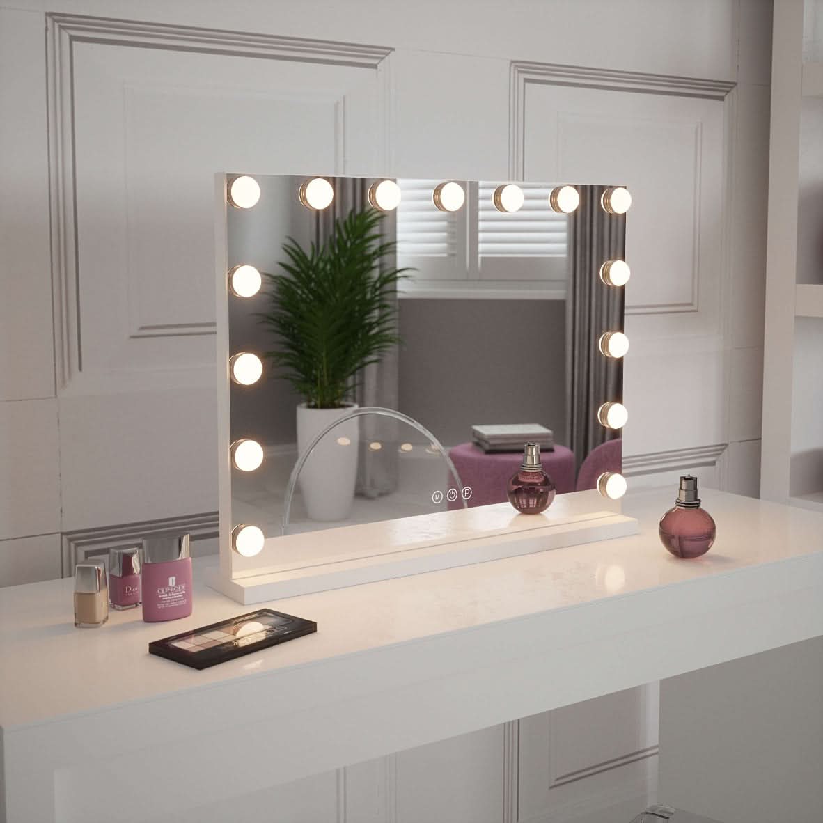 The Jessica Hollywood Mirror 58cmX46cm with LED bulbs sits on a white table, surrounded by perfume bottles, nail polish, and a makeup brush. Its versatile lighting reflects a green plant and window in the mirror’s glass.