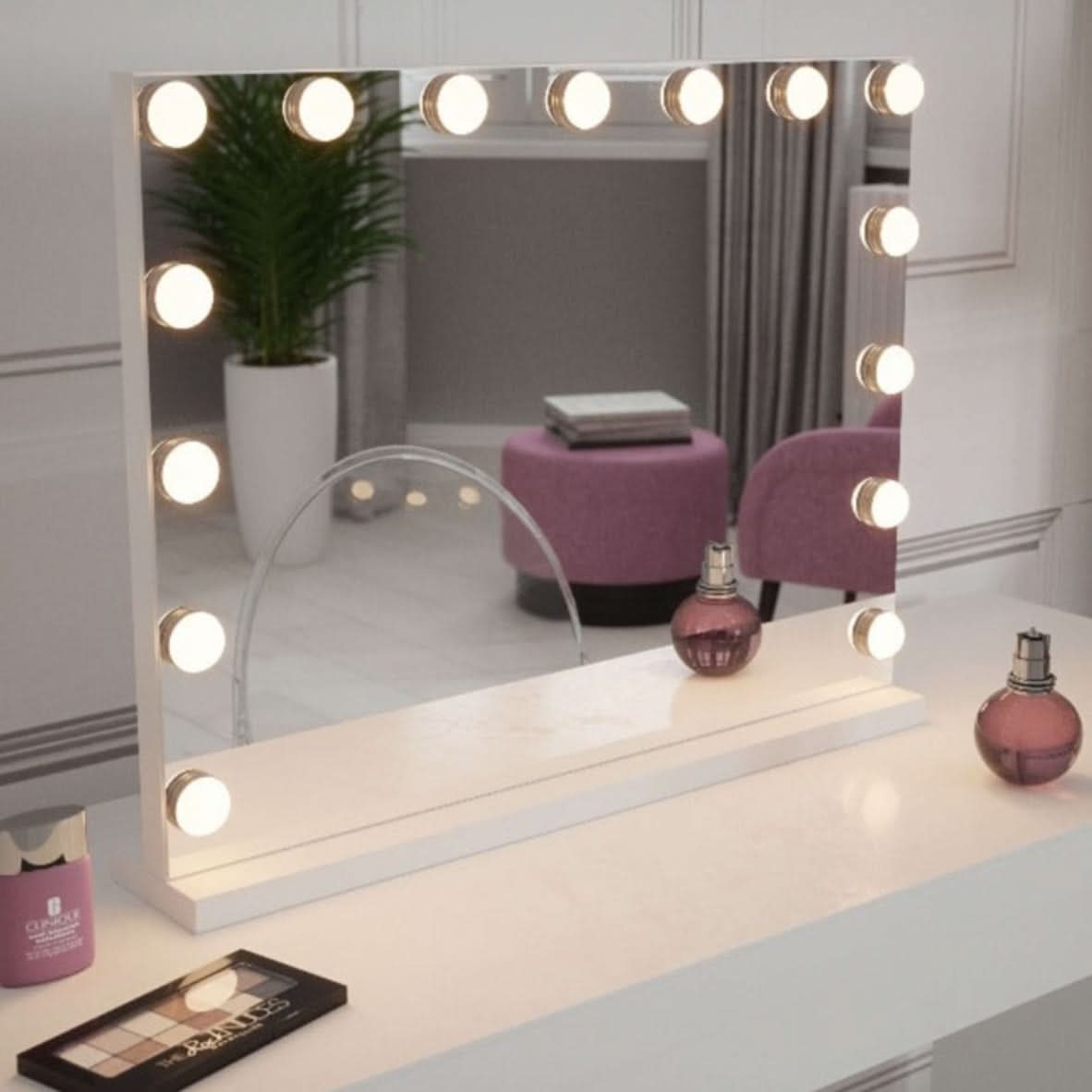 The Jessica Hollywood Mirror 58cmX46cm, with LED bulb lighting, sits on a dressing table among makeup and perfume. A clear chair, potted plant, and purple ottoman enhance the background style.