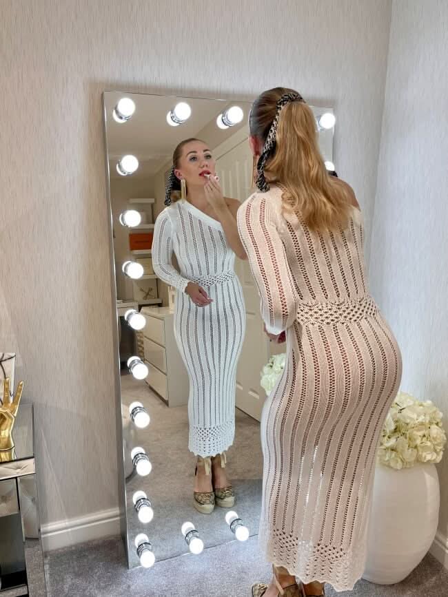 Woman applying makeup in front of XL Hollywood mirror with lights, featuring dimmer switch for perfect lighting, leaning against wall.