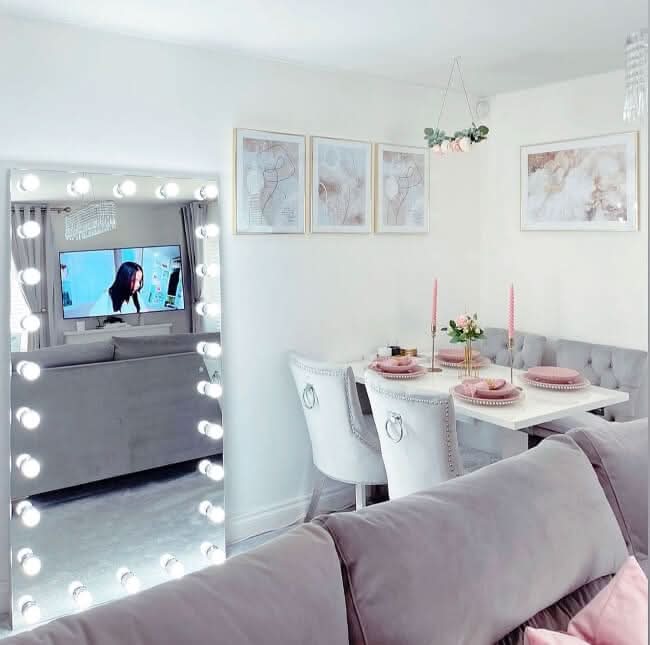 Chic dining room with Hollywood mirror showcasing bright white bulbs, pink table setting, gray couch, and wall art decor.