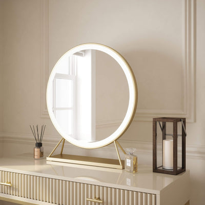 Anne Round Hollywood Mirror Gold 60x63cm with customizable lighting and touch sensor controls sits on a cream dresser, alongside a fragrance diffuser, perfume bottle, and lantern. A window is reflected in the mirror.