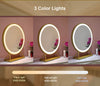 Three PRE ORDER Mid JUNE Anne Round Hollywood Mirrors Gold 60x63cm with gold stands are shown under warm, natural, and cold lighting. Each has customizable lighting, a touch sensor, and reflects a vase with pink flowers.