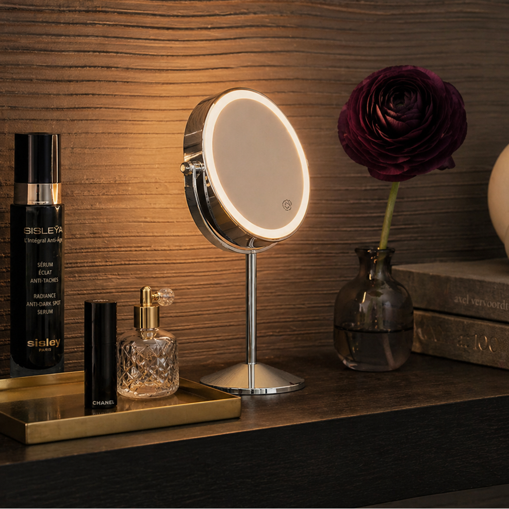 Small round mirror with lights on a wooden surface next to skincare products and a rose.