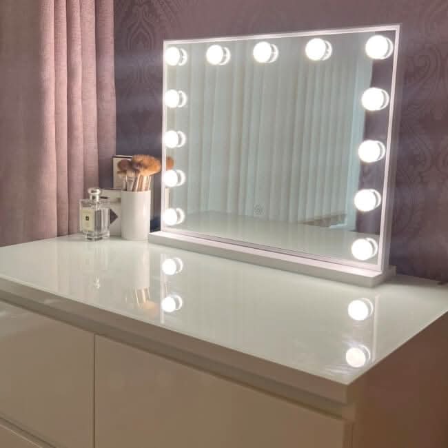 A white vanity table features the Margot Small Hollywood Mirror 50x42cm with adjustable pro lighting. To the left, a makeup brush holder and perfume bottle are set against curtains and patterned wallpaper.
