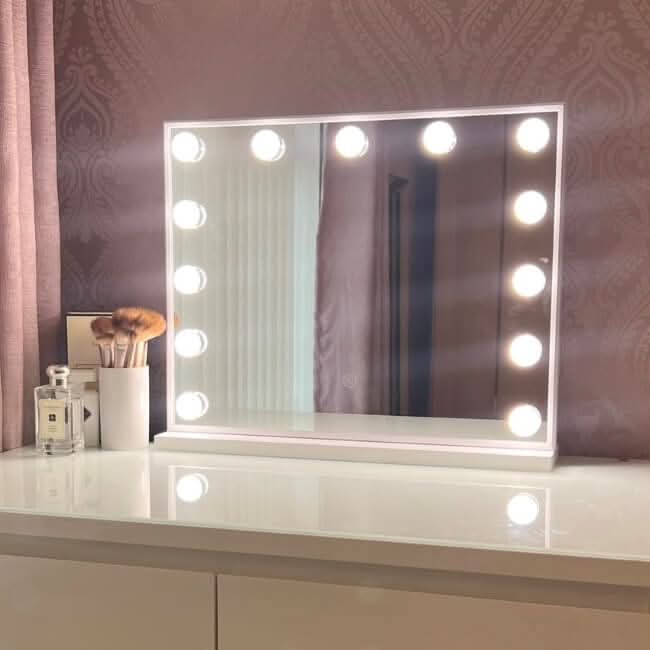 A vanity table with a Margot Small Hollywood Mirror 50x42cm featuring pro-grade lighting and adjustable brightness, makeup brushes in a holder, a perfume bottle, and soft patterned wallpaper in the background.