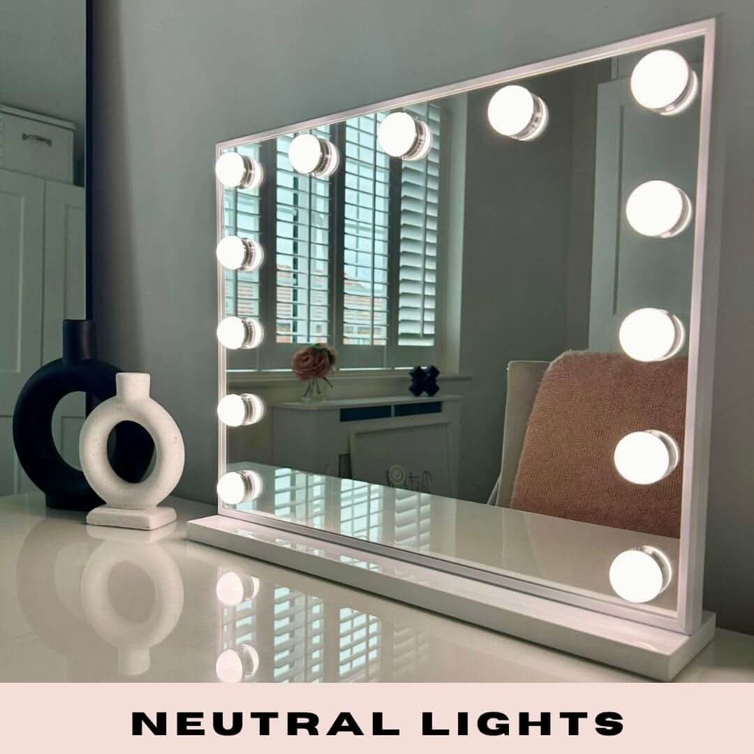 A Margot Small Hollywood Mirror 50x42cm with round neutral lights sits on a white table beside black and white vases. A chair and window blinds are in the background. Text reads: NEUTRAL LIGHTS.