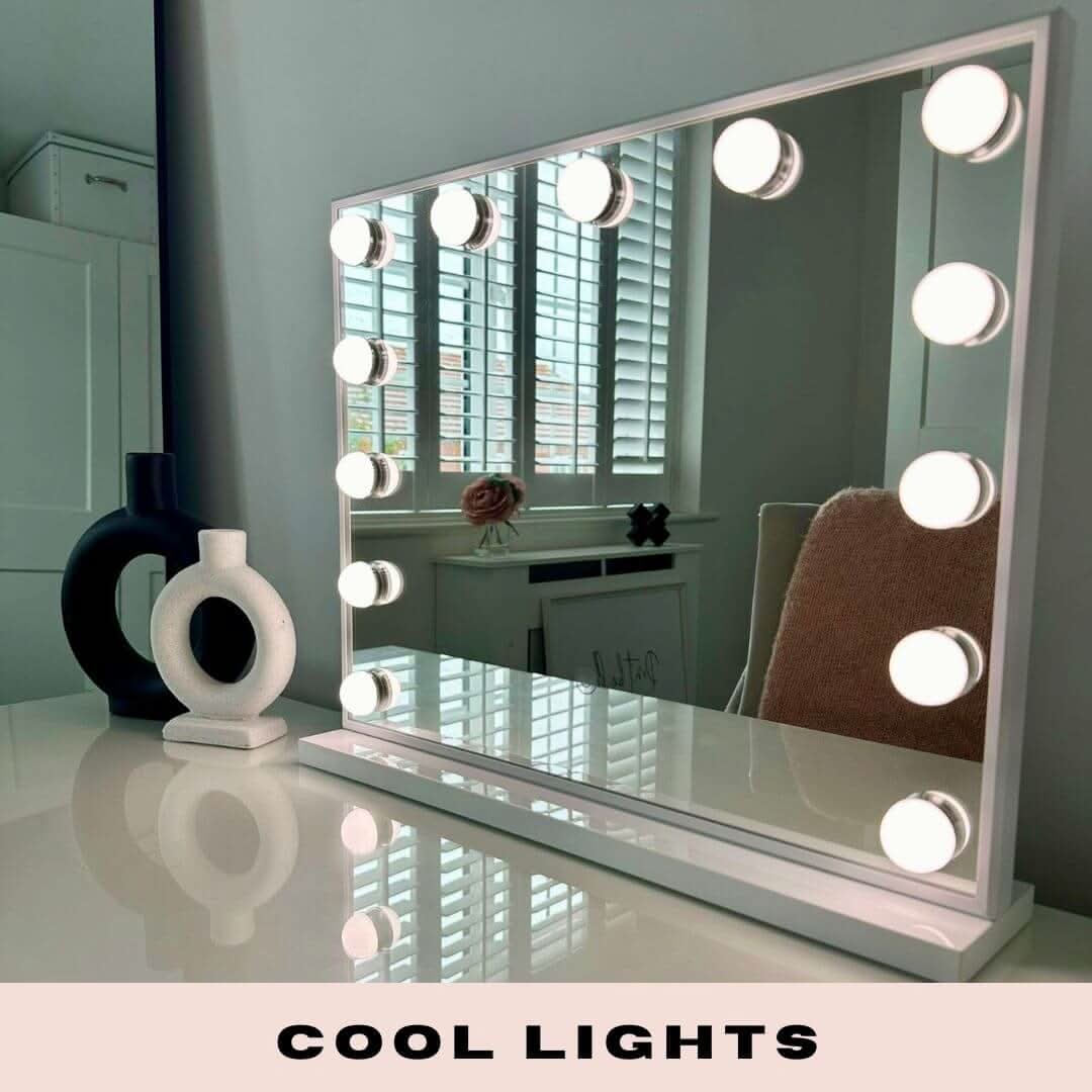 A Margot Small Hollywood Mirror 50x42cm, with round illuminated bulbs and adjustable brightness, sits on a glossy white desk decorated with vases. The mirror reflects a window with blinds. Text at the bottom reads: COOL LIGHTS.