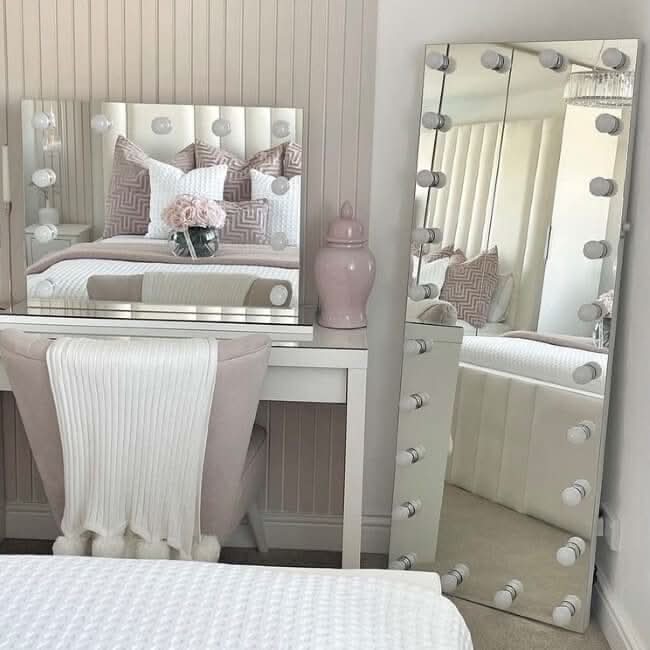 A chic vanity area showcases a white desk, cushioned chair, and the Mariah Full Length Hollywood Mirror 160x60cm by hollywood mirrors with round bulbs. A pink vase adds flair, while pink and white pillows on the bed are reflected in the elegant mirror.