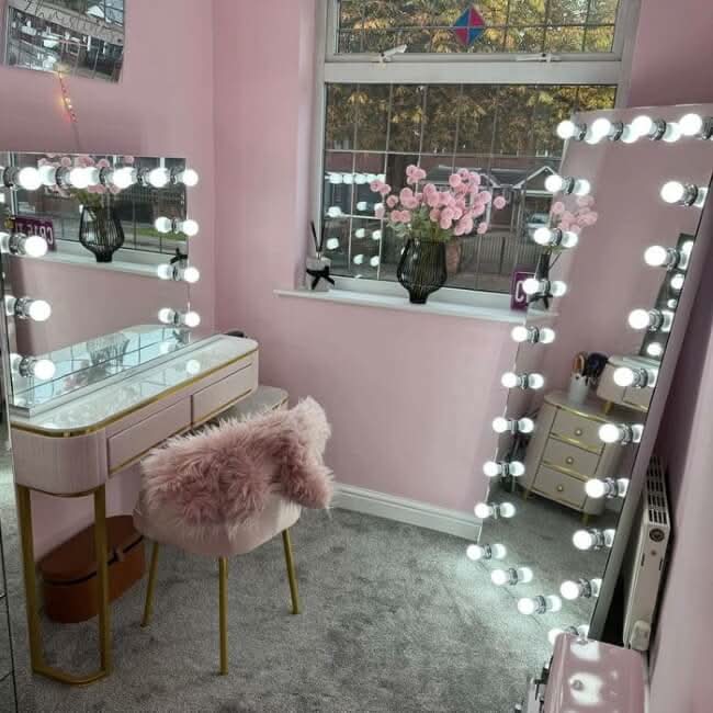 A pink-themed vanity area features the hollywood mirrors Mariah Full Length Hollywood Mirror 160x60cm, a fluffy pink chair, pink flowers on the window sill, a soft gray carpet, and warm light streaming through the window.