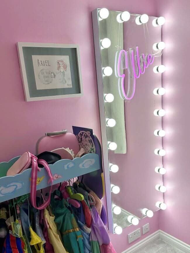 A pink room with a dress rack of colorful costumes, a neon Ellie sign, a framed Ariel drawing above the rack, and the Mariah Full Length Hollywood Mirror 160x60cm by hollywood mirrors framed with round lights.