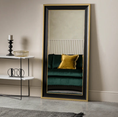 The Hudson Black & Gold Full Length Mirror by Hollywood Mirrors leans against the wall, reflecting a green sofa with a yellow pillow. A small white table nearby holds a candle, bowl, and books.