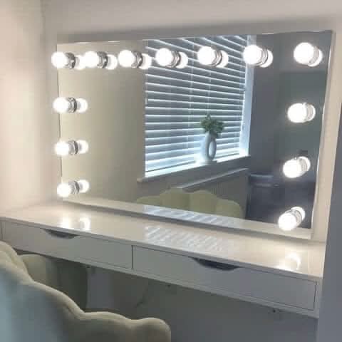 A white vanity table with drawers, a soft chair, and the hollywood mirrors Nicole Hollywood Mirror Wall Mounted 100x60cm featuring bright LED bulbs. The mirror reflects a window with closed blinds and a vase of green foliage.