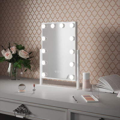 The Olsen Hollywood Mirror White 47x37cm by hollywood mirrors sits on a white vanity with adjustable LED bulbs, makeup items, a lipstick, flowers in a vase, and an open book against patterned wallpaper.