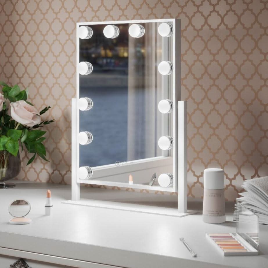 The Olsen Hollywood Mirror White 47x37cm by hollywood mirrors sits on a white desk with round LED bulbs, makeup items, roses, and an open book. Its adjustable brightness provides perfect lighting against beige wallpaper and a scenic window view.