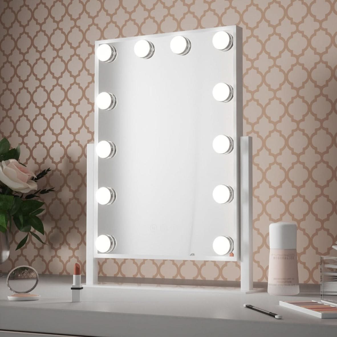 The Olsen Hollywood Mirror White 47x37cm by hollywood mirrors, featuring LED bulbs and adjustable brightness, sits on a desk with patterned wallpaper, alongside a flower vase, lipstick, jar, crystal ball, and closed makeup container.