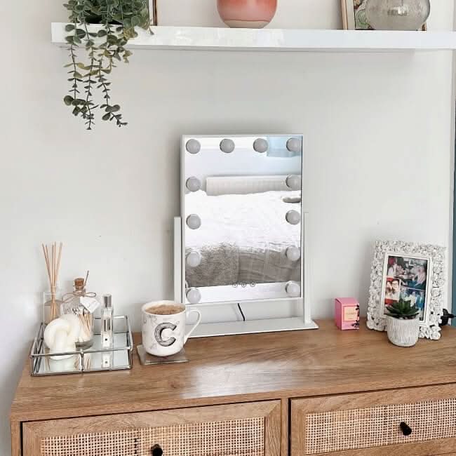A wooden dresser displays the hollywood mirrors Olsen Hollywood Mirror White 47x37cm with adjustable LED bulbs, a mug, candle, reed diffuser, and framed photo. A shelf above holds a potted plant and decor. The scene is bright and tidy.