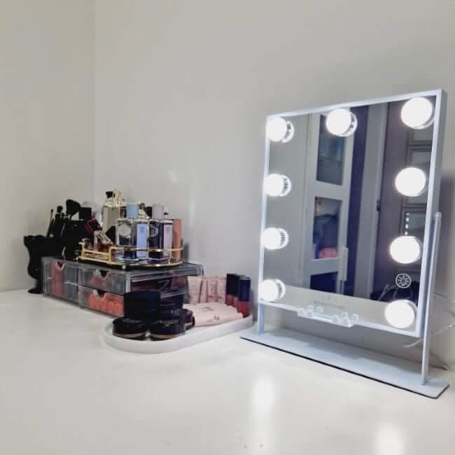 A vanity setup features the Renée Bluetooth Hollywood Mirror 30x25cm with LED bulbs, along with makeup brushes, perfumes, and assorted beauty products neatly arranged on a white tabletop.