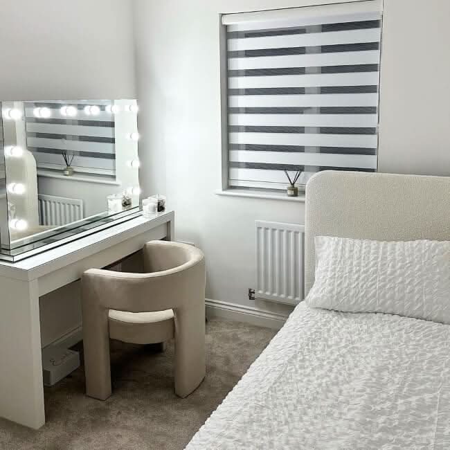 A modern, minimalist bedroom with a white bed, beige chair, and window with striped blinds features the hollywood mirrors Scarlett Large Hollywood Mirror 100x70cm on the vanity. Decor is mostly white and neutral tones.