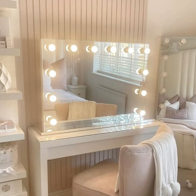 A cozy vanity setup features the hollywood mirrors Scarlett Large Hollywood Mirror 100x70cm with round LED bulbs, set against a beige paneled wall. A soft, light-colored chair draped with a blanket sits in front of the vanity.
