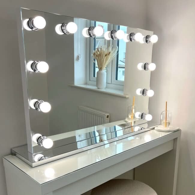 A hollywood mirrors Scarlett Large Hollywood Mirror 100x70cm is paired with a vanity table, cushioned stool, a windowsill vase with dried grass, and two drinks with straws placed on the tabletop.
