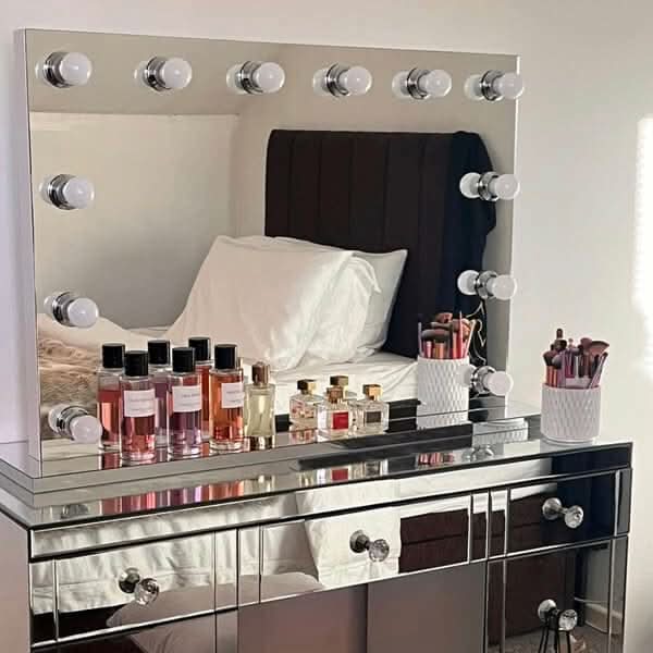 The Scarlett Large Hollywood Mirror 100x70cm by hollywood mirrors sits on a mirrored vanity table with LED bulbs, displaying perfume bottles, makeup brushes in a holder, and a white container. The mirror reflects a neatly made bed with white pillows.