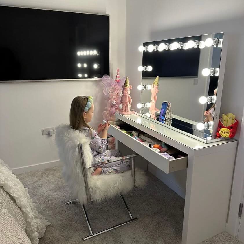 A young girl in pajamas applies makeup at a hollywood mirrors Scarlett Large Hollywood Mirror 100x70cm, surrounded by makeup, dolls, and a fries-shaped container. A large TV hangs nearby in the softly lit, cozy room.