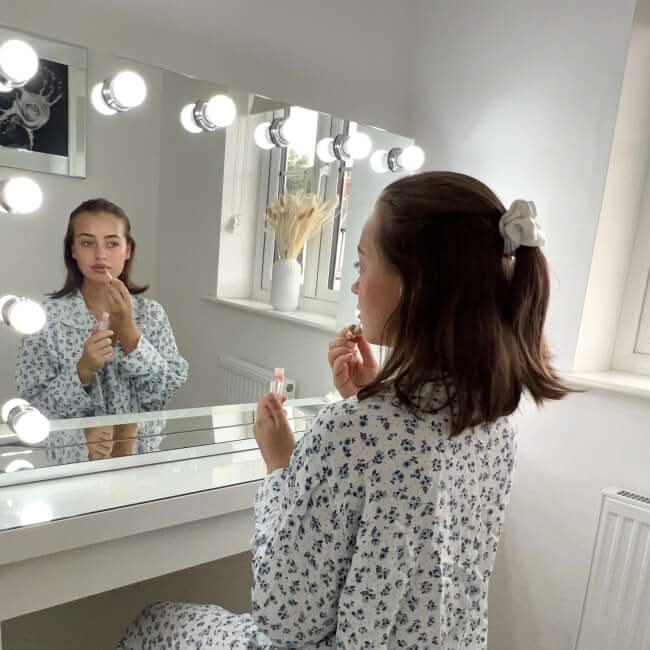 A woman with brown hair in a white floral robe applies lipstick while seated at the hollywood mirrors Scarlett Large Hollywood Mirror 100x70cm, featuring glowing LED bulbs in a bright, modern room.