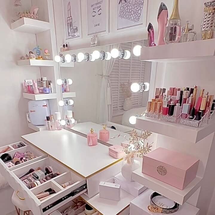 The Scarlett Large Hollywood Mirror 100x70cm by hollywood mirrors features LED bulbs and sits atop a white and pink vanity with organized makeup, skincare, and perfume neatly arranged on shelves and drawers in a stylish, bright room.