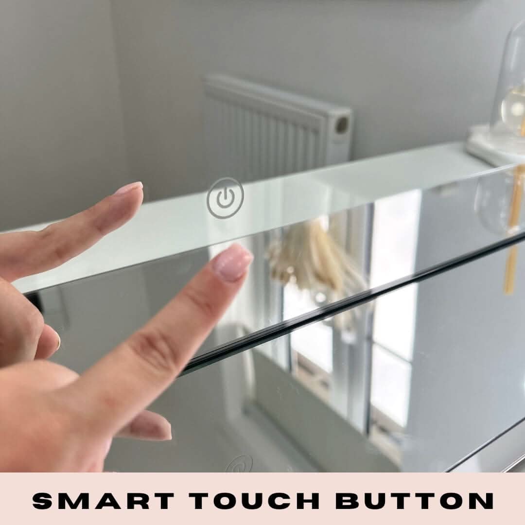 A person with manicured nails is about to tap the smart touch button of the Scarlett Large Hollywood Mirror 100x70cm by hollywood mirrors. LED bulbs softly illuminate the scene, with a radiator and window visible in the background.