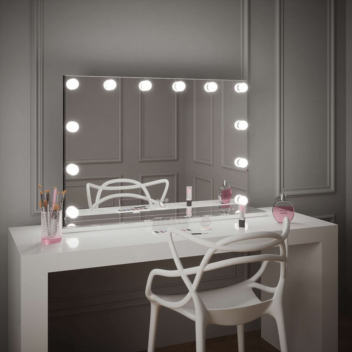 A Scarlett Large Hollywood Mirror 100x70cm by hollywood mirrors is mounted on a gray paneled wall above a white vanity table holding makeup brushes, perfume bottles, and cosmetics. A modern white chair sits in front.
