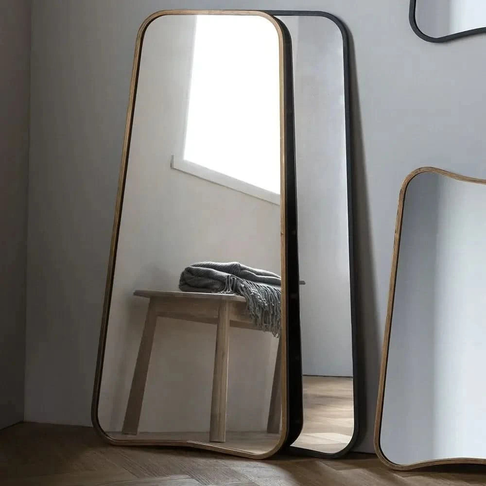 The hollywood mirrors Valentina Gold Mirror Full Length Mirror (123cm x 56.5cm) reflects a wooden bench with a gray blanket by a window in a softly lit room with warm wooden floors.