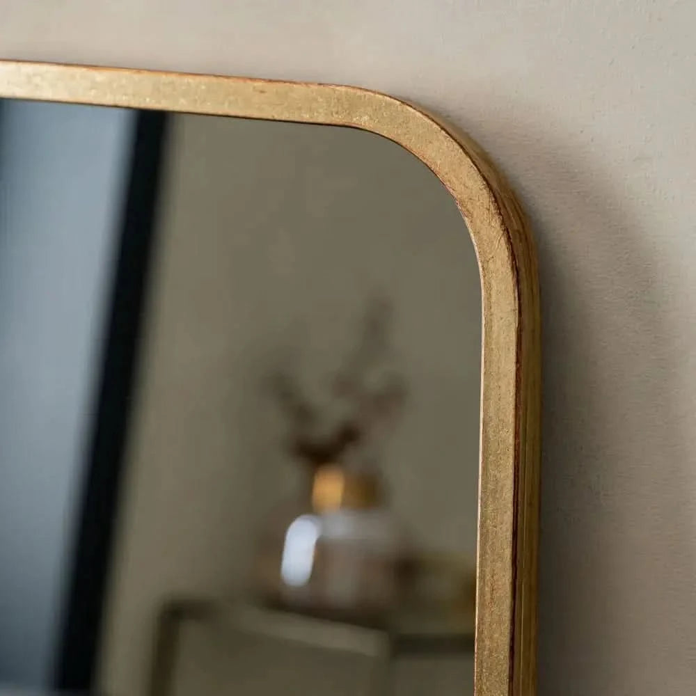 Close-up of The Valentina Gold Mirror Full Length Mirror by hollywood mirrors (123cm x 56.5cm), a rectangular design with a gold frame and rounded corners, reflecting a blurred decorative object against a neutral wall.