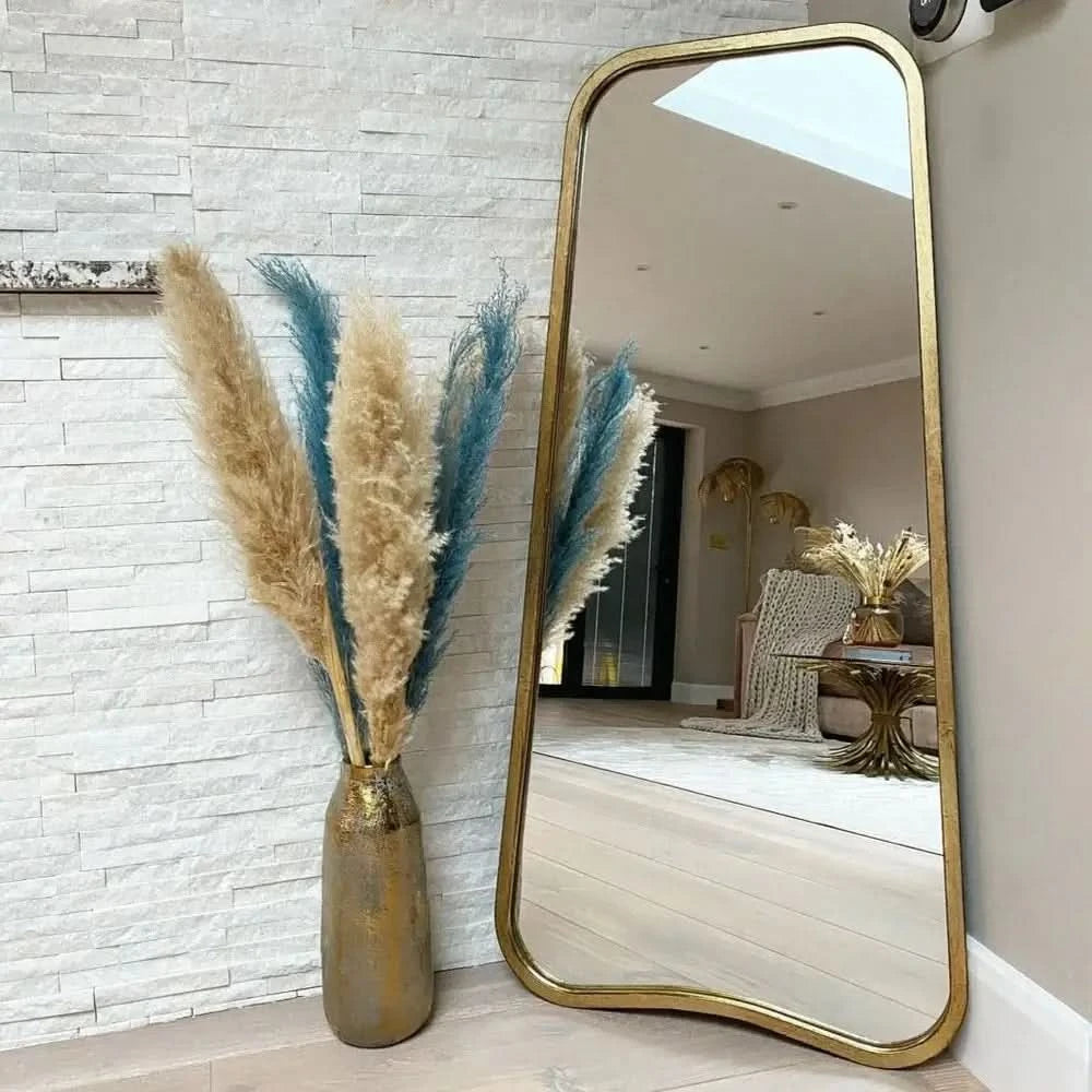 The hollywood mirrors Valentina Gold Mirror Full Length Mirror (123cm x 56.5cm) leans against a white textured wall next to a tall vase of beige and blue pampas grass, reflecting a stylish, modern living room.