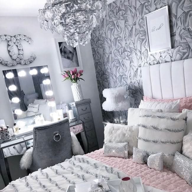 A chic, modern bedroom in pink, white, and silver features the hollywood mirrors Alicia Hollywood Mirror 60cmx80cm with LED bulbs and dimmer. Fluffy pillows, a crystal chandelier, framed art, and a bold Chanel logo complete the look.