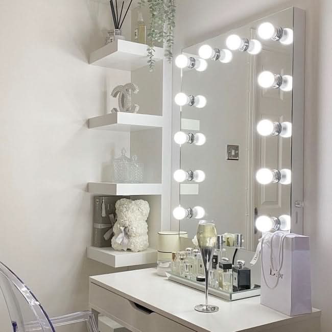 A champagne glass, perfumes, a white teddy bear, and a Dior gift bag sit on a vanity table with the Alicia Hollywood Mirror 60cmx80cm by hollywood mirrors; white wall shelves with decor and plants are beside the mirror.