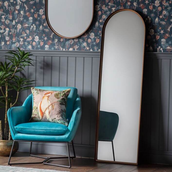 Bronze Arch Leaner Mirror 170x51cm in modern living room with blue chair, floral wallpaper, and wood paneling.