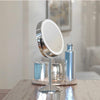 Illuminated Round Cosmetic Mirror Rechargeable, 20cm diameter, with an adjustable stand is on a wooden table beside a shiny silver bottle, metallic container, and decorative tray; a blurred painting is behind.