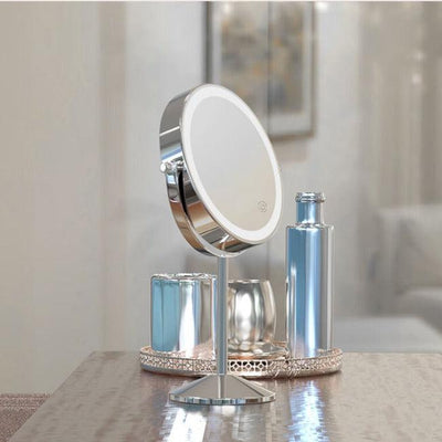 Illuminated Round Cosmetic Mirror Rechargeable, 20cm diameter, with an adjustable stand is on a wooden table beside a shiny silver bottle, metallic container, and decorative tray; a blurred painting is behind.