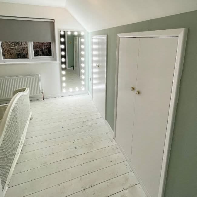 A bright, minimalist bedroom with white floors, a bedframe, gray blinds over the window above the radiator, a built-in white closet, and a hollywood mirrors Mariah Full Length Hollywood Mirror 160x60cm surrounded by lights.