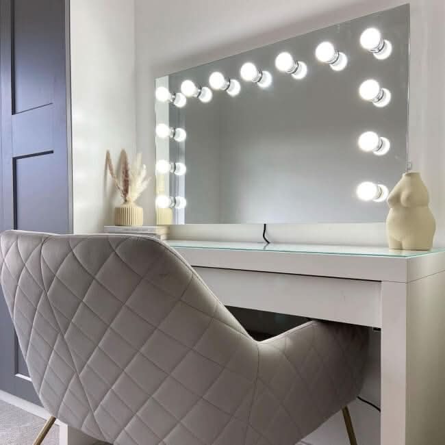 A vanity setup featuring the Nicole Hollywood Mirror Wall Mounted 100x60cm by hollywood mirrors, a white desk, a quilted gray chair, plus beige pampas grass in a vase and a decorative beige figure on the tabletop.