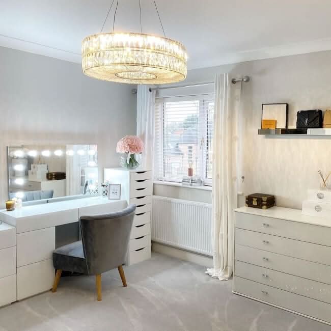 A bright, modern vanity room features the hollywood mirrors Nicole Hollywood Mirror Wall Mounted 100x60cm with LED bulbs, a gray chair, white drawers, shelves with decor, pink flowers, and sunlight streaming through white-curtained windows.