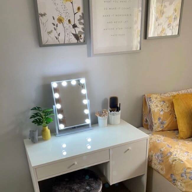 A white vanity table with the hollywood mirrors Olsen Hollywood Mirror White 47x37cm featuring adjustable brightness sits beside a floral yellow bed, adorned with potted plant, brushes, and decor. Framed botanical prints hang on the light gray wall above.
