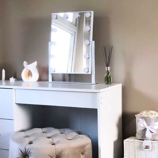 A white vanity table with the hollywood mirrors Olsen Hollywood Mirror White 47x37cm with adjustable brightness, a tufted gray stool, cat-shaped candle holder, reed diffuser, and ribboned white basket, arranged against a beige wall.